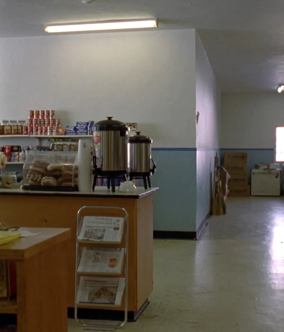 The inside of a small country convenience store before opening, with a laminate counter, two stainless coffee urns, a stack of paper cups, a small glass donut case and shelves behind under fluorescent light.