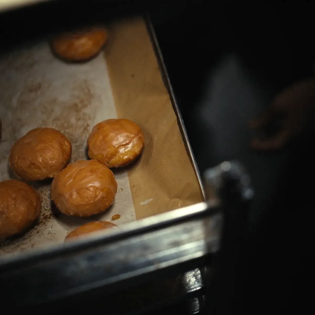 A tray of plain glazed cake donuts cooling on wax paper on a steel rack, uneven and handmade, lit warmly from one side.