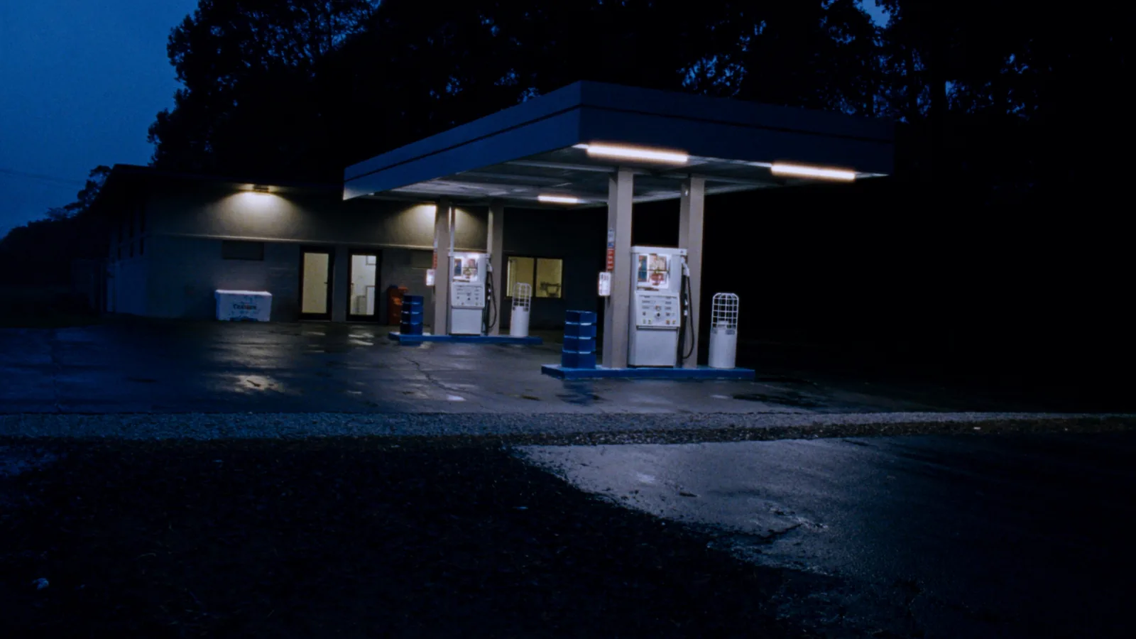 A small rural gas station an hour before sunrise, with two fuel pump islands under a lit metal canopy, a low block building with warm lit windows behind, and wet asphalt reflecting the lights.