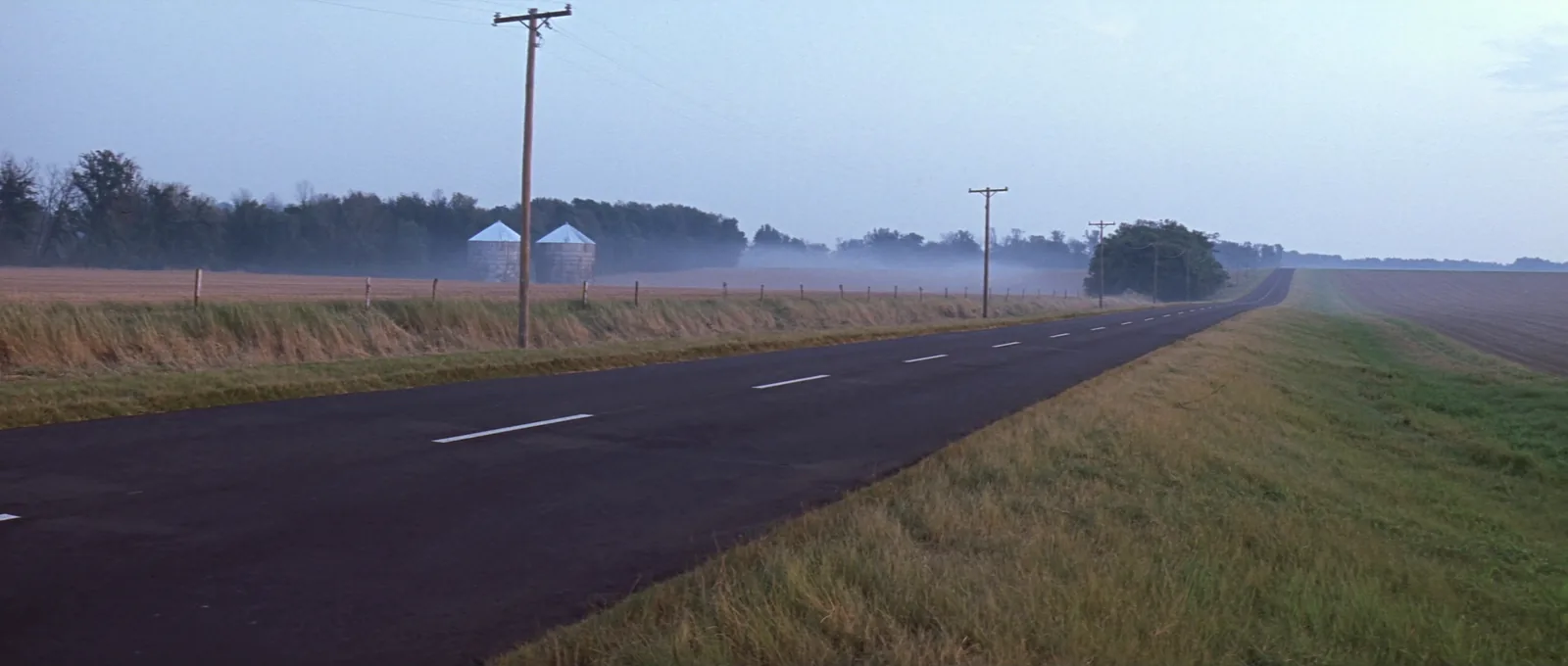 A two lane blacktop road running straight through Missouri farmland at dawn, with low mist over the fields, a line of power poles and two grain bins in the distance.